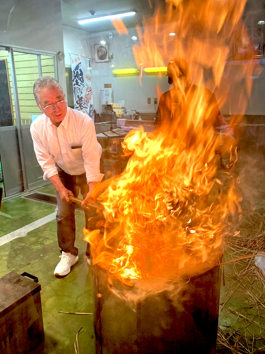 「焼津さかなセンター」(静岡県焼津市)で鰹藁焼き体験。焼津港に冷凍してある鰹一本を川直のお兄さんが捌いてくれて、その生の鰹を藁を燃やした炎でこんがり焼いて鰹のタタキを作ります。その焼いているところの写真です。焼いている人は岩本町三丁目町会の町会員、男性Xさんです。2026年6月28日、岩本町三丁目町会の親睦旅行での一幕です。