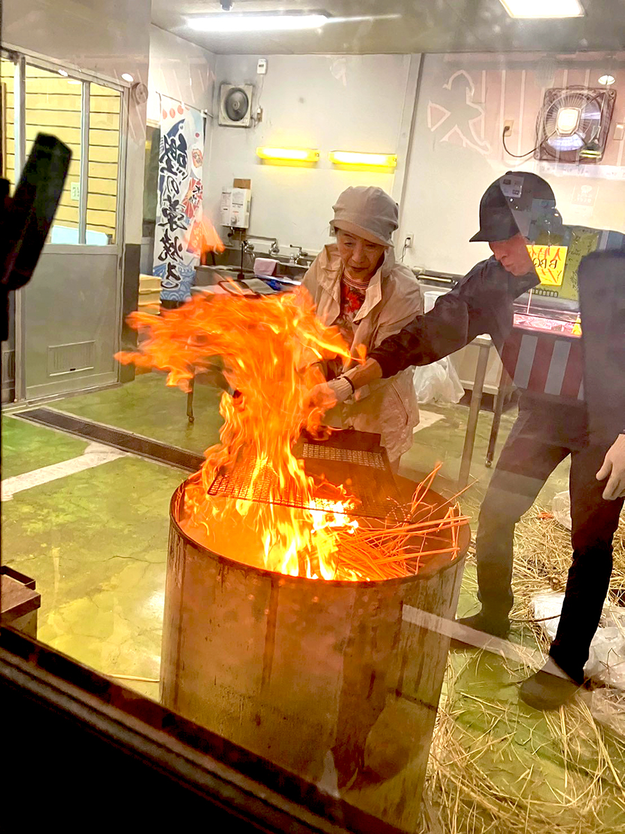 「焼津さかなセンター」(静岡県焼津市)で鰹藁焼き体験。焼津港に冷凍してある鰹一本を川直のお兄さんが捌いてくれて、その生の鰹を藁を燃やした炎でこんがり焼いて鰹のタタキを作ります。その焼いているところの写真です。焼いている人は岩本町三丁目町会の町会員、女性Yさんです。一緒に写っているのが川直のお兄さんです。2026年6月28日、岩本町三丁目町会の親睦旅行での一幕です。