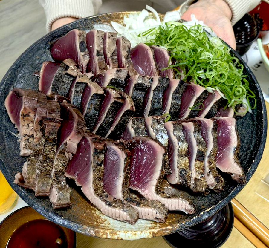 「焼津さかなセンター」(静岡県焼津市)で鰹藁焼き体験で、岩本町三丁目町会の皆さんが焼いた鰹のタタキです。茶色の陶器の丸い大皿に分厚い切り身がずらっと並んでいます。マイナス60度冷凍の解凍したてだから鮮度良好。綺麗な生の部分の外側の焼けた部分が「岩三」のみなさんが手を加えたところです。おいしかったです。2026年6月28日、岩本町三丁目町会の親睦旅行での一幕です。