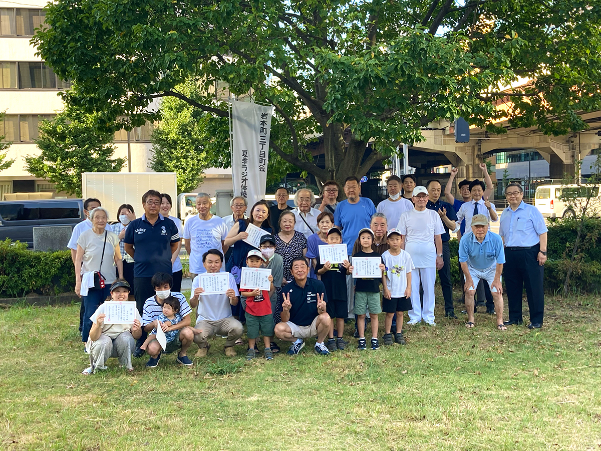 2023年8月3日、岩本町三丁目町会の夏季ラジオ体操、最終日の集合写真です。全日参加されたみなさんが賞状を手ににこやかに写真に写っています。小さなお子さん、お父さんお母さんもにっこり。ラジオ体操の会場は「馬の水飲広場」（うまのみずのみひろば。東京都千代田区3-10　岩本町交差点内）です。交差点内なのに木も生えていて、公園のような広い草地で。この、都心では珍しいオアシスのゾーンで木を背景にして、草地に全員集合して記念写真をパチリと撮りました。岩本町三丁目町会では毎年7月下旬から8月上旬にかけて夏季ラジオ体操をおこなっていて、この写真は岩本町三丁目町会の町会新聞『リバーサイドイワモト３』33号（2024年正月発行）から特別に転載したものです。