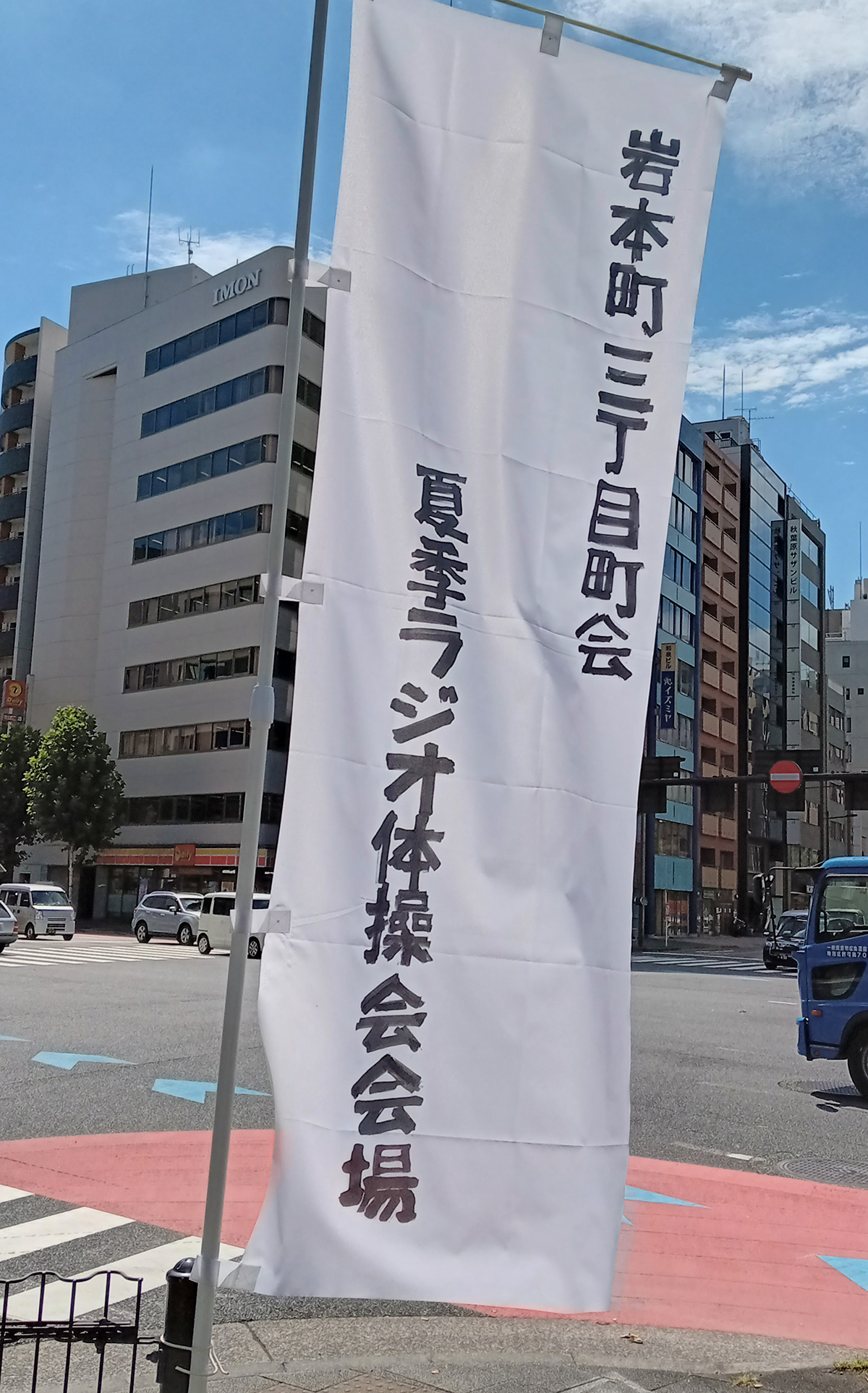 2023年から岩本町三丁目町会の夏季ラジオ体操の会場は、大門通りから「馬の水飲広場」（うまのみずのみひろば。東京都千代田区3-10）へ変更となりました。昭和通り、靖国通り、水天宮通りの交わる緑のゾーン。大きな木がある公園のような広い草地でラジオ体操をおこなうようになったのです。この会場変更を機に夏季ラジオ体操開催期間限定で「馬の水飲広場」にラジオ体操の会場だと示す幟を立てることにしました。この写真はその幟です。幟の旗竿をプラスチック製の白。幟の旗地は純白。そこに黒のフェルトペンで「岩本町三丁目町会、夏季ラジオ体操会」と手書き。アットフォームな幟です。その幟が夏の風に気持ちよくたなびいています。幟の背景には靖国通りと水天宮通りが写っています。この写真は岩本町三丁目町会の町会新聞『リバーサイドイワモト３』33号（2024年正月発行）から特別に転載したものです。