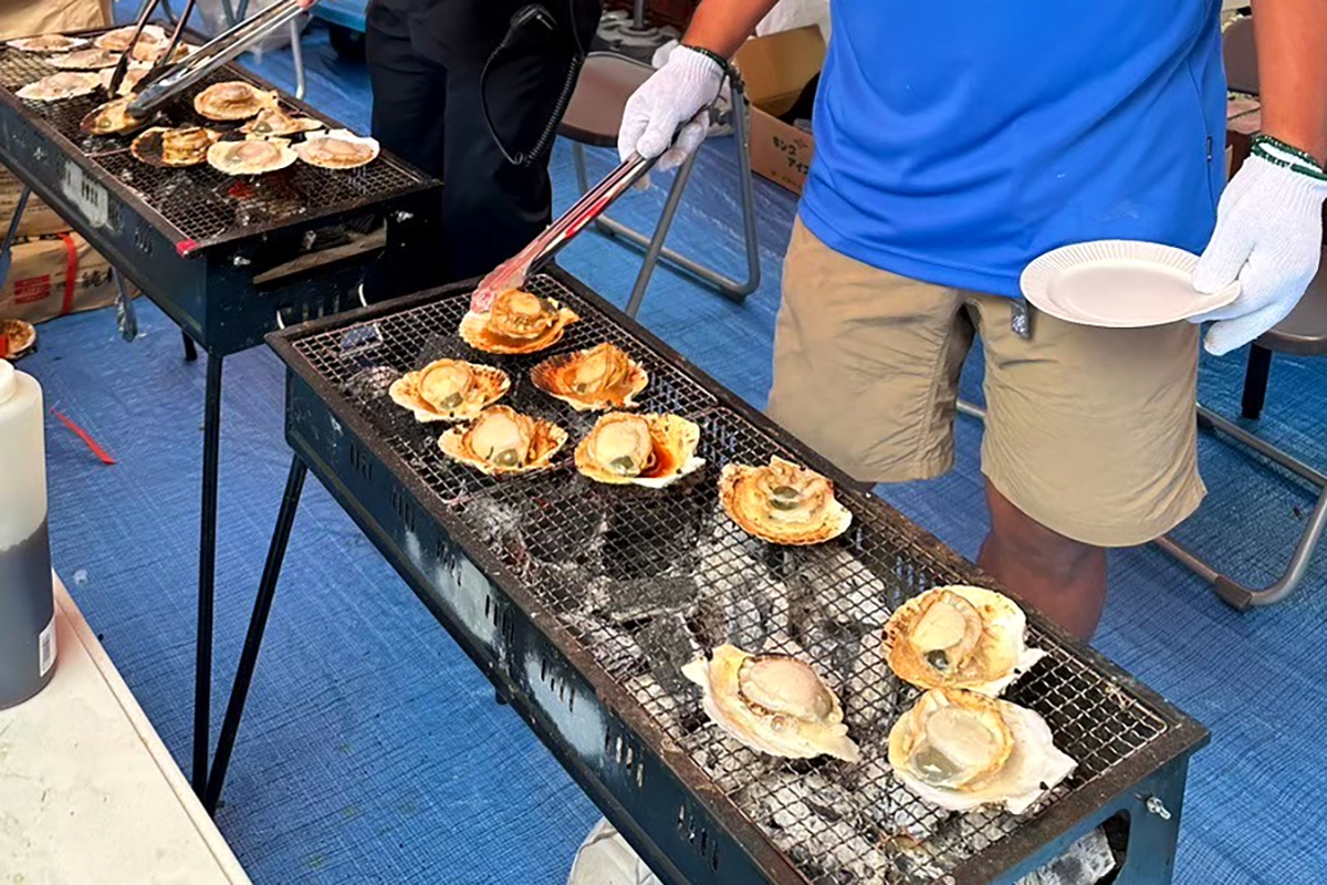 岩三名物「ほたて焼き」の写真です。刺身にできる新鮮なホタテ貝を炭火でじっくり焼き上げています。帆立貝の殻の深い方をお皿代わりにして、その上に帆立の身を乗せて、焼き網の上に並べて焼いているところです。手前の貝にご注目。貝柱以外の部分（例　ウロやヒモ）が付いたまま新鮮でしっかり下処理しているから、こういう風に食べられます。奥の貝は出来上がり直前。岩三秘伝のタレがかかった程よい焼き上がりに仕上がっています。岩本町東神田町会連合会の納涼祭で食べられます。岩本町三丁目町会自慢の一品に舌鼓を打ってください。