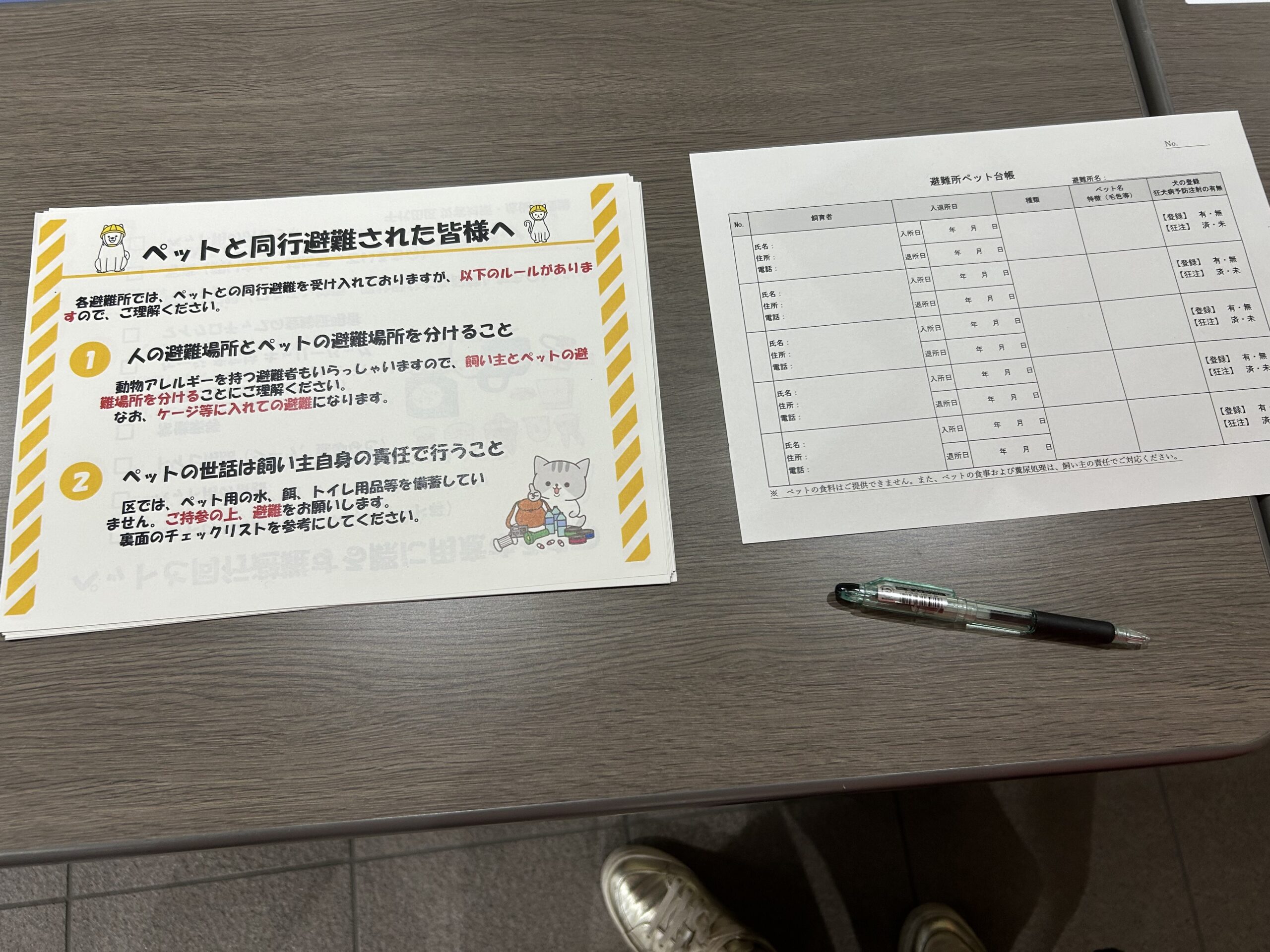 災害時に神田岩本町そして岩本町三丁目等の住民のために開設される「岩本町ほほえみプラザ」避難所の開設訓練で設置されたペット避難の受付の写真です。受付の机の上にペット避難のルール（ペットと人は別の場所で＝「同行避難」）を書いた紙と、ペット情報の記入票（インフルエンザ注射の有無などを記入）が置いてあります。